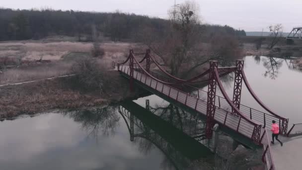 Egy nő fut át egyedül a hídon, légi kilátással. Női kocogás reggel a parkban a folyóval. Egy lány fényes sportruhában átfut a hídon a folyón, drónlövéssel. — Stock videók