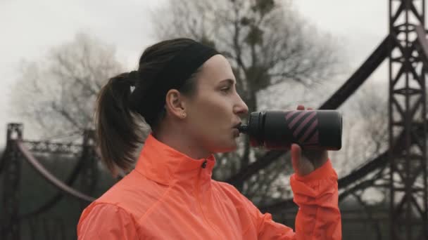 Portret młodej kobiety pijącej napój energetyczny z butelki. Brunetka sportowiec pije wodę z butelki na świeżym powietrzu w parku. Dziewczyna w pomarańczowej kurtce przygotowuje się do treningu. Koncepcja sportu — Wideo stockowe