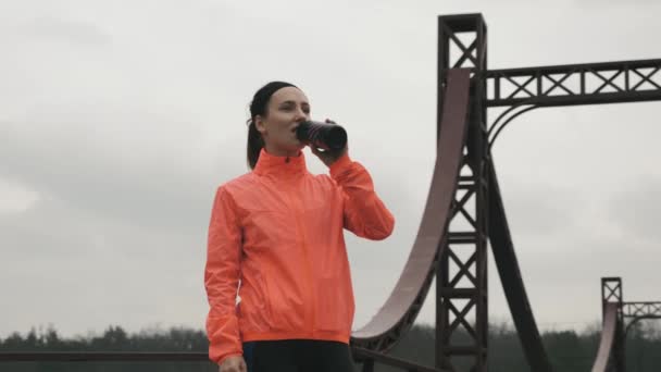 Female athlete is drinking water outdoor in park. Young sportive woman in bright sportswear drinks energy drink from bottle before training. Sports and running concept — Stock Video