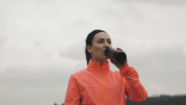 Brunett kvinnlig idrottsman dricksvatten från flaska i parken. Ung kvinna i ljusa sportkläder drycker från flaska utomhus. Idrott och löpning — Stockvideo