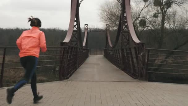 La donna con le cuffie sta attraversando il ponte nel parco. Giovane sportiva in giacca arancione e collant neri corre sul ponte attraverso il fiume all'aperto. Sport e stile di vita sano concetto — Video Stock