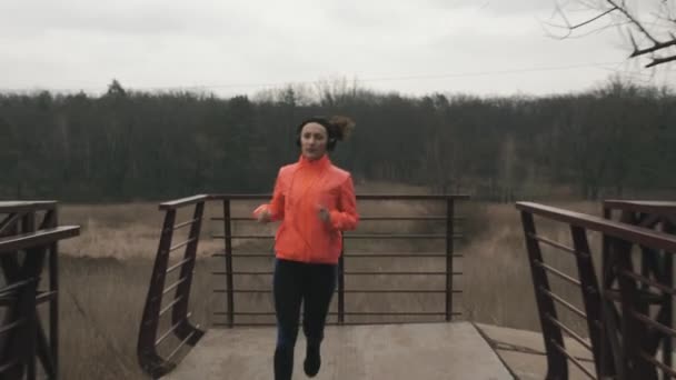 Morena atleta feminina em fones de ouvido que atravessa a ponte pedonal em parque. Jovem mulher em jaqueta laranja corre na ponte através do rio na área de recreação. Menina fazendo exercícios matinais — Vídeo de Stock