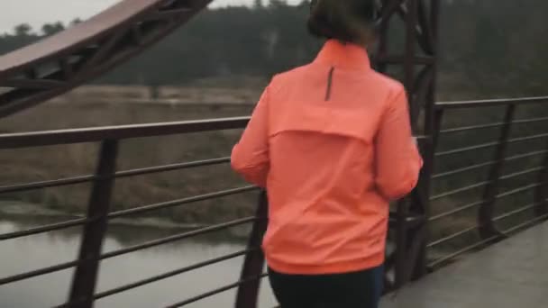 Kvinnan joggar utomhus. Ung kvinnlig idrottsman i ljusa sportkläder som löper på bron i parken. Sport och kör koncept — Stockvideo