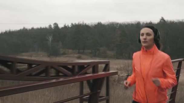 El retrato de una joven con auriculares cruza el puente. Morena atleta femenina en chaqueta naranja trotando al aire libre en el parque. Formación de deportistas en zonas verdes — Vídeos de Stock