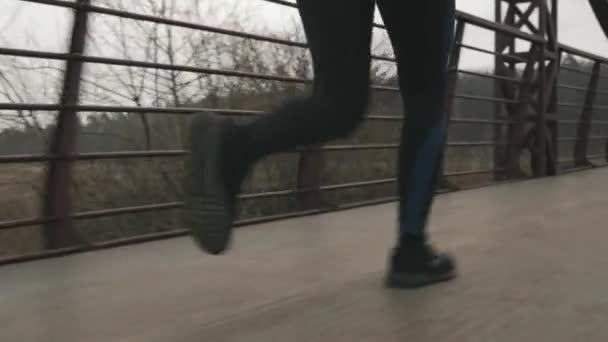 Kvinnliga ben springer utomhus, närbild bakåt. Kvinnliga fötter joggar i parken på bron över floden. Idrott och löpning — Stockvideo