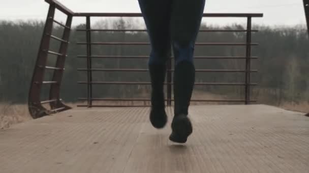 Vista frontal de las piernas de la mujer corriendo en el puente. Pies femeninos en mallas negras cruzan el puente en el parque. Concepto de deporte y running — Vídeos de Stock