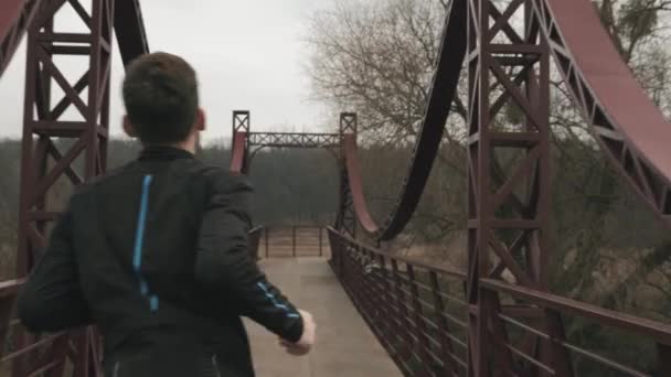 Athlète masculin sportif traversant le pont en plein air. Homme caucasien en vêtements de sport noirs jogging sur le pont à travers la rivière dans le parc. Guy formation dans la zone de loisirs — Video