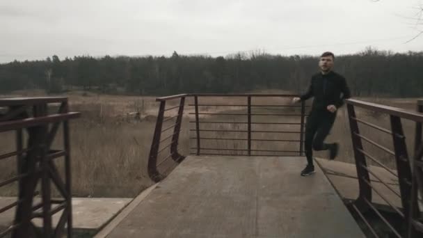 Man springer på bron över floden, framifrån. Vit ung man i sportkläder som springer utomhus i parken på gråmulen dag. Tunn kille utbildning i skog med flod och bro — Stockvideo