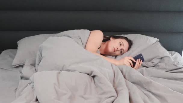 갈색 머리 여자가 침대에 누워서 스마트폰 화면을 보고 있습니다. 외로운 젊은 여성 이 손에 전화기를 들고 혼자 침대에 누워 있다 — 비디오