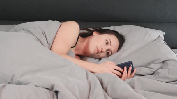 Portret młodej dziewczyny leżącej na szarym łóżku i piszącej na smartfonie. Brunetka leży na pustym łóżku i patrzy na smartfona. Urocza kobieta z telefonem w rękach leżąca sama w łóżku. — Wideo stockowe