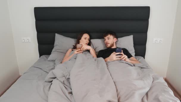 Mujer y hombre acostados en la cama con teléfonos inteligentes en las manos. Joven pareja casada en cama gris viendo dispositivos y mensajes de texto en las redes. Pareja femenina y masculina en el dormitorio — Vídeos de Stock