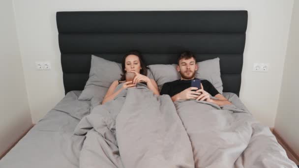 Kvinnor och män ligger i sovrummet och tittar på sina smartphones. Kris i förhållande. Svårigheter i förhållandet. Unga gifta par tittar på smartphones och chatta på internet i sängen — Stockvideo