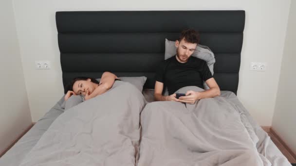 Jeune homme trichant sur sa femme en ligne. Couple marié couché au lit. Le mari envoie des SMS en ligne par téléphone portable tandis que sa femme est allongée sur le bord du lit seule. Problème sexuel. Crise dans les relations — Video