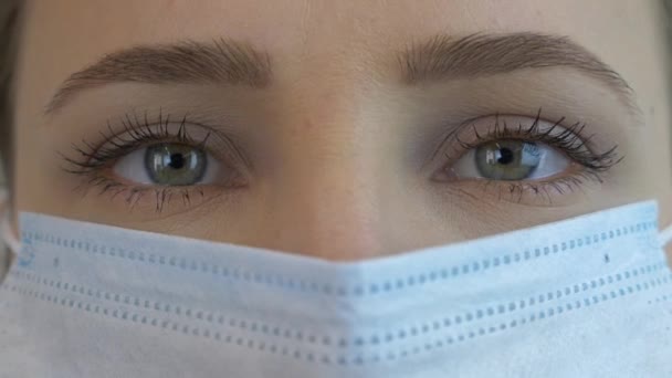 Θηλυκό πρόσωπο σε ιατρική μάσκα προσώπου για την πρόληψη και την προστασία των λοιμώξεων του ιού, εξαιρετικά κοντά. Πανδημική προστασία του Covid-19 coronavirus. Γυναίκα με πράσινα μάτια σε προστατευτική ιατρική μάσκα — Αρχείο Βίντεο
