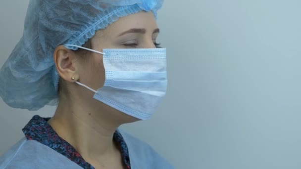 Portret van een vrouw die een beschermend medisch gezichtsmasker afdoet. Jonge vrouwelijke wetenschapper doet een medisch blauw masker af. Dokter verwijdert beschermend gezichtsmasker na chirurgische behandeling — Stockvideo