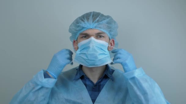 Мужчина-врач в защитном снаряжении надевает медицинскую маску перед операционным лечением. Человек в защитной медицинской одежде готовится к операции в клинике. Пандемическая защита коронавируса — стоковое видео