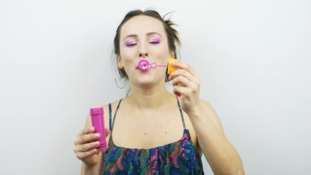 Porträtt av ung glad flicka blåser såpa bubblor mot vit bakgrund. Kvinna med rosa makeup har roligt. Ung brunett kvinna blåser såpbubblor — Stockvideo