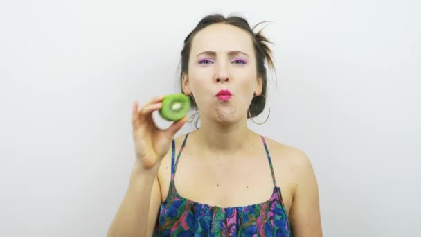 Portret van een lachend meisje dat groene kiwi eet tegen een witte achtergrond. Jonge vrolijke vrouw eet kiwi en kijkt naar de camera. Vrouw heeft plezier — Stockvideo