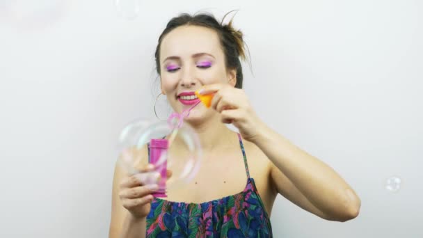 Счастливая смеющаяся девушка с розовым макияжем, дующая мыльными пузырями. Смешная улыбающаяся женщина дует мыльные пузыри на белом фоне. Женщина развлекается. — стоковое видео