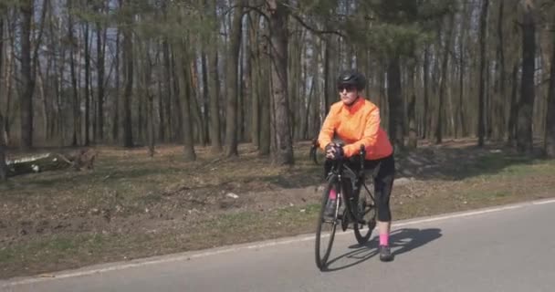 Nő biciklin, portré. Egy nő áll biciklivel az erdőben az üres úton. Kerékpáros sportoló sisakban, felkészülve a biciklis edzésre. Kerékpározás és triatlon koncepció — Stock videók