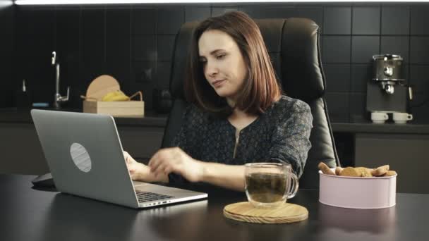 年轻的女自由职业者在家里的笔记本电脑上工作时头疼。在厨房的笔记本上工作的女人很不高兴.不幸的女性在家里使用计算机技术。可怜的女孩看着手提电脑屏幕 — 图库视频影像