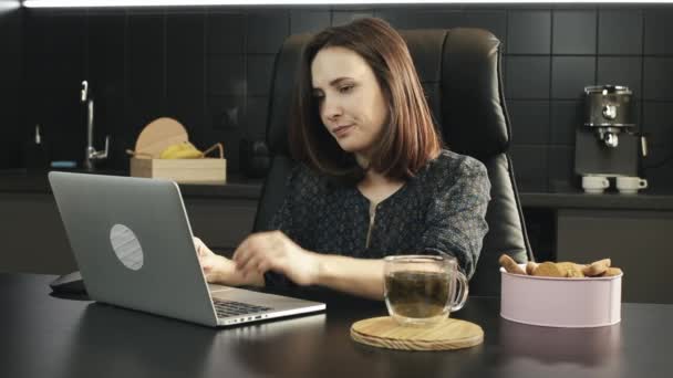 Joven freelancer con dolor de cabeza trabajando en la computadora portátil en casa. Mujer molesta trabajando en un cuaderno en la cocina. Las mujeres infelices usan la tecnología informática en casa. Chica triste mirando a la pantalla del ordenador portátil — Vídeos de Stock