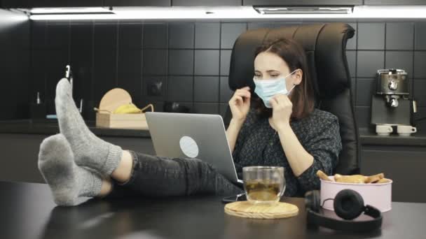 Mladá žena si nasadí ochrannou masku a začne doma pracovat na notebooku. Žena na volné noze pracující v ochranné masce obličeje z domova během karantény — Stock video