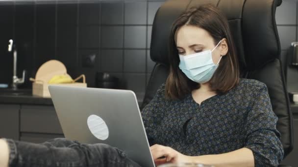 Vrouw die een beschermend medisch gezichtsmasker gebruikt terwijl ze thuis aan een laptop werkt. Meisje met medisch masker typt op schrift in de keuken thuis. Portret van vrouwelijke freelancer in het gezicht masker werk vanuit huis — Stockvideo