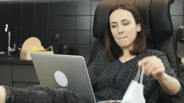 Vrouw zet beschermende medische masker voordat online werk op laptop computer thuis werkplek. Een vrouw die thuis online werkt. Persoon met een medisch gezichtsmasker werkt op schrift in de keuken — Stockvideo