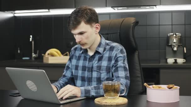 Freelancer masculino usando laptop en casa. Hombre caucásico con bigote beber té durante el trabajo en línea en el ordenador portátil en la cocina. Persona que trabaja desde casa y escribe en el teclado del ordenador portátil — Vídeos de Stock
