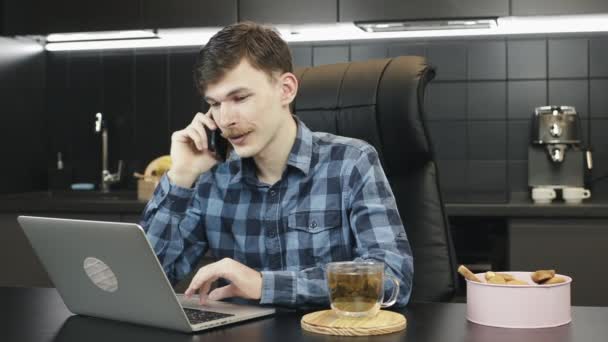 Um homem a fazer um telefonema amigável de casa. Jovem freelancer masculino trabalhando computador portátil a partir de casa e falando no smartphone. Empresário conversando com contato comercial ou amigo no local de trabalho — Vídeo de Stock