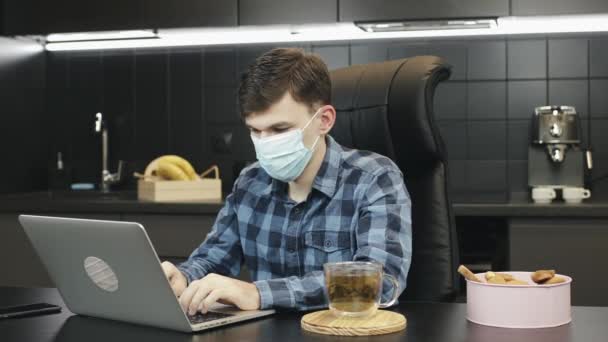Человек, использующий защитную медицинскую маску для лица во время работы над ноутбуком дома. Мужчина в медицинской маске, печатает на блокноте дома на кухне. Портрет мужчины-фрилансера в маске для лица работы из дома — стоковое видео