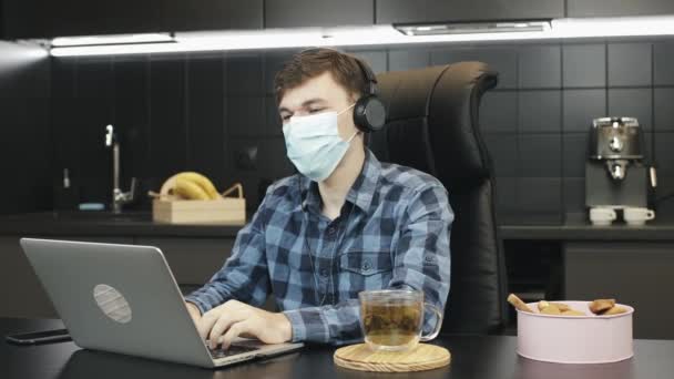 自宅の職場からビデオ通話会議を作る保護医療面の男。自宅で保護顔マスクを身に着けている人。自宅のラップトップで作業中に医療用マスクを使用して男性 — ストック動画