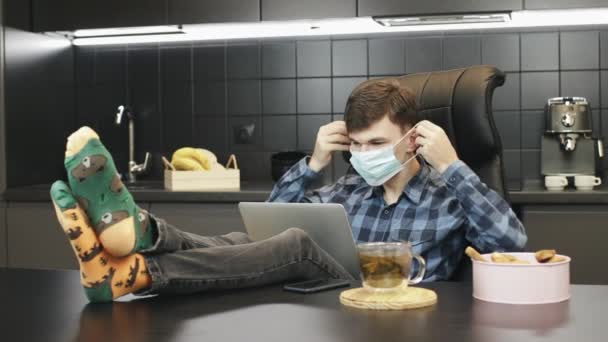 Az ember védőmaszkot vesz fel a járvány megelőzésére, és otthon elkezd dolgozni a laptopon. Orvosi maszkban dolgozó férfi otthonról a karantén alatt — Stock videók