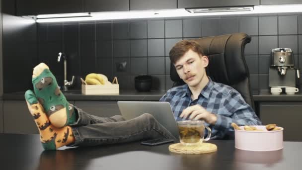 キッチンに座っている男は、自宅でラップトップコンピュータでリラックスしてオンラインで作業しています。ノートを使って自宅のオフィスで働く若い男性。キーボードを入力してノートパソコンの画面を見る白人男性 — ストック動画