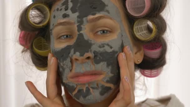 Kobieta w czarnej, glinianej masce na twarzy patrząca w kamerę, z bliska. Kobieta z lokami na głowie ma zabiegi kosmetyczne w domu. Pielęgnacja skóry i naturalne pojęcie kosmetyczne. Kosmetologia w domu — Wideo stockowe