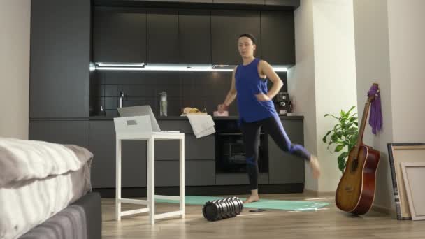 Mujer fitness haciendo ejercicio y ejercicios aeróbicos en casa. Entrenamiento femenino en casa en línea utilizando la aplicación en el ordenador portátil. Ajuste chica haciendo ejercicios de la mañana en la cocina luminosa en casa. Deporte en casa — Vídeos de Stock