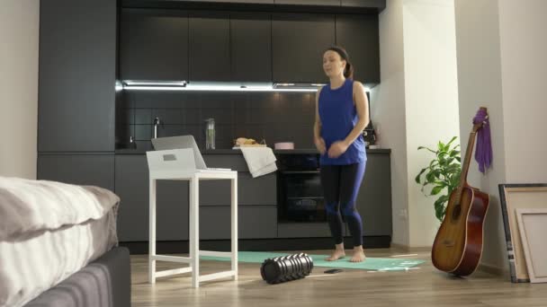 Mujer saltando y haciendo ejercicios aeróbicos de fitness en casa en la cocina utilizando la aplicación de entrenamiento en línea en el ordenador portátil. Ajuste chica deportiva en ropa deportiva atlética haciendo ejercicios de deporte por la mañana en casa — Vídeos de Stock