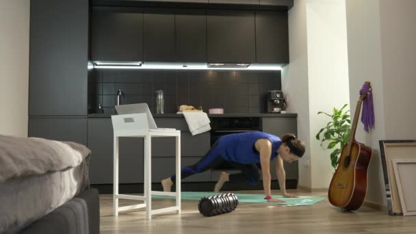 ラップトップコンピュータ上のオンライントレーニングアプリを使用して台所で板を練習する強いフィットネスの女の子。自宅で女性の訓練。スポーツウェアを着た女性が家で働いている — ストック動画