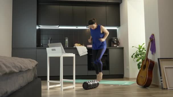 Mulher atlética forte energeticamente correndo e fazendo exercícios de fitness on-line em casa na cozinha. Treinamento feminino em casa usando o aplicativo no computador portátil. Menina fazendo exercícios matinais em casa — Vídeo de Stock