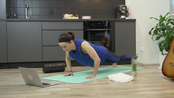 Chica haciendo ejercicios aeróbicos y de entrenamiento en línea en casa para un estilo de vida saludable. Ajuste mujer en ropa deportiva haciendo ejercicios de fitness utilizando la aplicación en línea en el ordenador portátil. Mujer hace ejercicios push up en la cocina — Vídeos de Stock