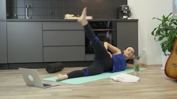 Женщина делает онлайн аэробные упражнения на тренажерном коврике дома с помощью приложения на ноутбуке. Спортивная девочка тренируется дома во время карантина коронавируса. Женщина делает спортивные упражнения, оставаясь дома — стоковое видео