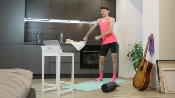 Homem de fitness engraçado estúpido dançando gostando de música e aquecendo para o treino na cozinha em casa. Atleta masculino engraçado com um bigode no topo rosa fazendo exercícios aeróbicos em casa — Vídeo de Stock