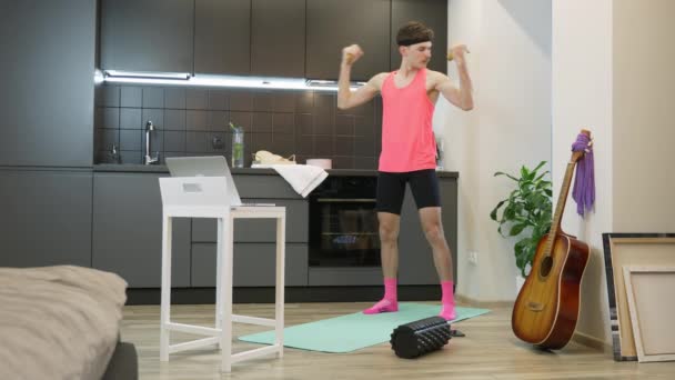Divertido tipo de aspecto estúpido en traje rosa haciendo ejercicios de entrenamiento en bíceps con plátanos en casa. Atleta masculino expresivo viendo clases de fitness en línea y haciendo ejercicios aeróbicos en casa en la cocina — Vídeos de Stock