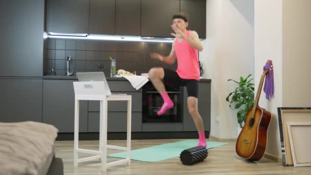 Joven atleta expresivo entrenar en casa y ver clases de fitness en línea en el ordenador portátil. Hombre divertido con bigote y traje rosa haciendo ejercicios aeróbicos en la cocina. Inicio entrenamiento deportivo — Vídeos de Stock