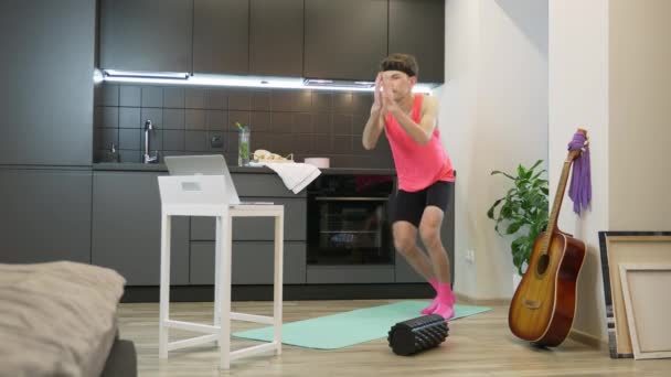 Chico joven haciendo ejercicios aeróbicos y de fitness en casa utilizando la aplicación de entrenamiento en línea en el ordenador portátil. Hombre divertido con bigote y top rosa de los 80 trabajando en la cocina quedándose en casa durante la cuarentena — Vídeo de stock