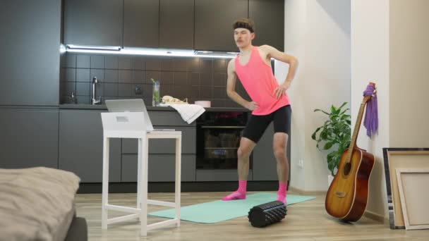 Смішний фітнес хлопець з вусами і рожевим вбранням дивиться онлайн-аеробні заняття на ноутбуці комп'ютера і робить фізичні вправи вдома. Молодий чоловік тренується вдома на кухні під час карантину — стокове відео