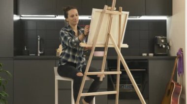 Modern mutfaktaki bar sandalyesinde oturan akrilik boya kullanarak tuvalde resim yapan kadın sanatçı. Kadın fırça kullanarak resim çiziyor. İlham. Sanat terapisi. Çizim okulu