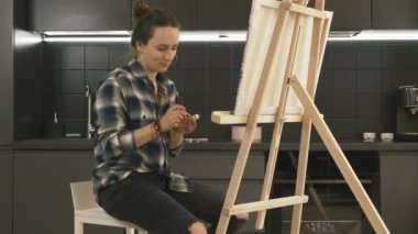 Kadın sanatçı, arka planda mutfak olan tuvalde resim yapıyor. Şövalenin yanındaki sandalyede oturan yetenekli genç kadın evde akrilik boya kullanarak resim çiziyor.