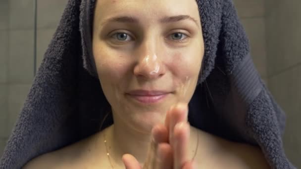 La mujer con toalla en la cabeza se aplica crema para el cuidado de la piel en la cara y se mira en el espejo después de la ducha. Mujer poner crema facial lifting hidratante. Concepto de salud y tratamiento — Vídeos de Stock